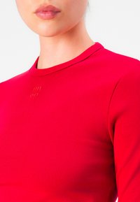 Close-up of a person wearing a bright red fitted shirt with a small embossed logo centered below the neckline.