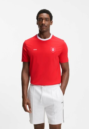 Man met een rood T-shirt met korte mouwen, een witte kraag en witte korte broek, staand tegen een lichte, effen achtergrond.