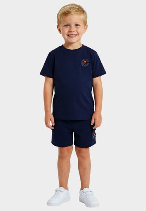 Jeune garçon souriant portant un t-shirt bleu marine et un short avec des logos de marques, debout devant un fond clair uni.