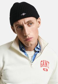 Zwarte ribgebreide beanie met wit logo, gecombineerd met een crèmekleurige zip-up sweatshirt met een rode graphic, gedragen over een blauw gestreept shirt.