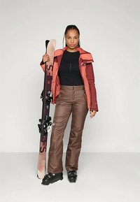 Σακάκι σκι σε κόκκινο και μπορντώ χρώμα, μαύρη μπλούζα, καφέ παντελόνι και μαύρες μπότες σκι. Κρατάει ένα σκι με δέστρες και λογότυπο.