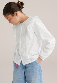 Blouse blanche à pois texturés, avec des volants le long des manches et une bordure en dentelle sur le devant, portée avec un jean bleu clair.