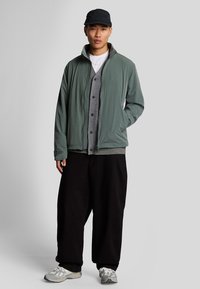 Ljusgrön zip-up-jacka över en grå cardigan, svarta vida byxor och grå sneakers. Jackan har en upphöjd logotypdetalj.