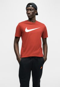 Νεαρός άνδρας που φοράει κόκκινο t-shirt Nike, μαύρη φόρμα Nike και μαύρο καπέλο με φορά προς τα πίσω, στέκεται με τα χέρια στις τσέπες.