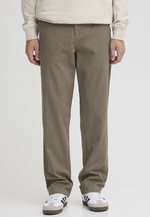 Mann trägt beige gerade geschnittene Hosen, weiße Turnschuhe mit schwarzen Streifen und ein cremefarbenes Langarmshirt, steht vor einem einfarbigen Hintergrund.