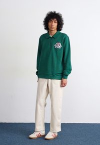 Grønn genser med krage og brodert logo, kombinert med kremfargede bukser og hvite sneakers med røde striper. Glatt stoff med løs passform.