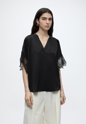 Femme aux longs cheveux foncés portant un haut noir ample avec des manches bordées de dentelle et un pantalon blanc à jambes larges sur un fond clair uni.