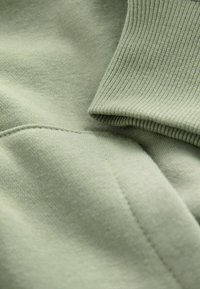 Hellgrüner Stoff mit glatten und gerippten Texturen, der Nähte und Falten zeigt – ideal für Freizeitkleidung oder zum Layering.