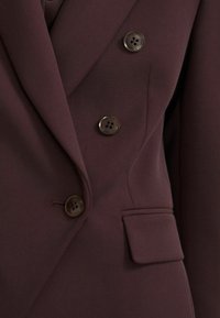 Bordo spalvos švarkas su dvigubu užsegimu, keturiais dideliais rudo atspalvio mygtukais, V formos atvartais ir viena kišene su atvartėliu. Lygios tekstūros.