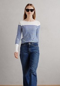 Kék és fehér csíkos, hosszú ujjú póló, hajónyakkal, magas derekú sötétkék farmerrel párosítva. A modellen oversize fekete napszemüveg van.