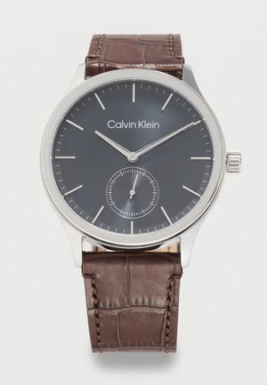 Ceas de mână Calvin Klein cu cadran negru, marcaje argintii, subcadran pentru secunde mici, carcasă argintie și curea din piele maro cu textură.