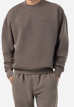 Hombre con sudadera de cuello redondo y pantalones deportivos a juego en color topo, con las manos en los bolsillos, de pie frente a un fondo liso.