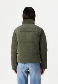 Olijfgroene corduroy jas met voering, hoge kraag en geribbelde textuur, met een boxy pasvorm. Draag het over blauwe spijkerbroeken.