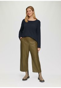 Vrouw staat gekleed in een zwarte langemouwtop, olijfgroene wijde broek en beige enkelboots met zwarte elastische zijkanten.
