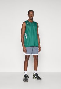 Zelený bezrúkový športový top z priedušného materiálu s bielymi bočnými pruhmi, skombinovaný so sivými šortkami s bielym lemom. Čierne športové topánky s neónovými akcentmi.