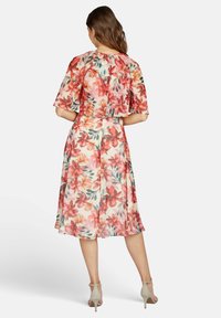Frau trägt ein knielanges Blumenkleid mit roten und pinken Blumen, steht mit dem Rücken zur Kamera und trägt beigefarbene High Heels.