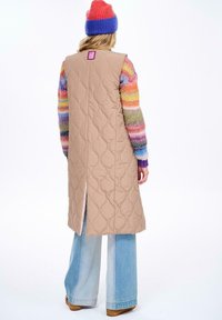 Beżowa, pikowana długa kamizelka z rozcięciem z tyłu, noszona na wielokolorowy sweter w paski. Posiada kwadratową metkę z tyłu.