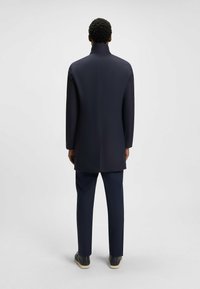 Cappotto lungo di un blu navy scuro con colletto alto, tessuto morbido e vestibilità dritta, abbinato a pantaloni coordinati e calzature semplici.