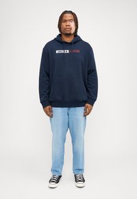 Felpa con cappuccio blu navy con tasca frontale, branding bianco e rosso, abbinata a jeans in denim azzurro chiaro e sneakers nere con suola bianca.