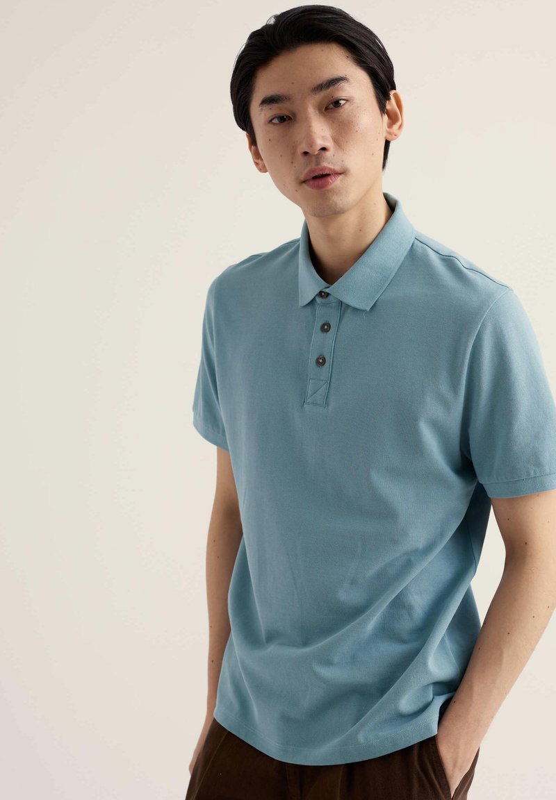 Jeune homme aux cheveux noirs portant un polo bleu clair à manches courtes, debout devant un fond beige uni.