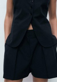 Gilet noir sur mesure avec un devant à six boutons et des poches latérales, associé à un short plissé assorti. Le tissu semble lisse et structuré.