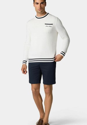 Witte gebreide trui met marineblauwe strepen op de kraag en manchetten, voorzien van een borstzak. Gecombineerd met marineblauwe shorts en bruine schoenen.