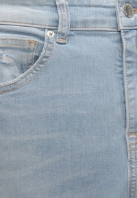 Ljust blå denimtyg med en framficka med synlig sömnad och silverknapp, vilket visar en blekt textur och revor.