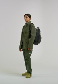 Giacca e pantaloni verde oliva in tessuto con multiple tasche. Zaino nero con finiture dorate. Scarpe color cuoio, in piedi su uno sfondo neutro.