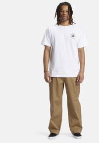 Hvid bomulds t-shirt med rund hals og et lille sort logo på venstre bryst, parret med sandfarvede lige ben bukser og sorte sneakers.