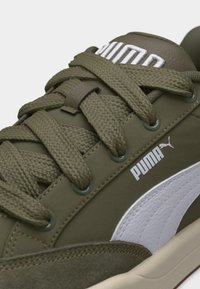 Gros plan d'une basket Puma vert olive montrant des lacets épais, des œillets en métal et le logo Puma blanc sur le panneau latéral texturé.