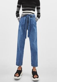 Niebieskie dżinsowe spodnie z wysokim stanem i paskiem do związania, o prostych nogawkach lekko zwężających się przy kostkach; noszone z czarnymi sandałami.