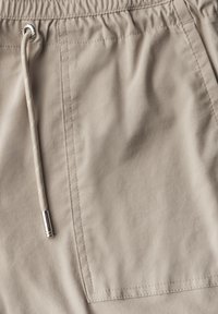 Pantaloni corti beige realizzati in un tessuto strutturato, con vita elasticizzata e cordoncino, e una tasca a toppa sul lato, decorata con cuciture sottili.