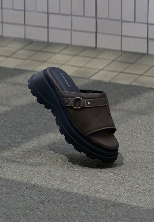 Braune Leder-Slipper-Sandale mit dicker schwarzer, robuster Sohle und Metall-Schnallen-Detail, die auf strukturiertem Pflaster liegt.