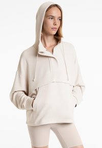 Sweat à capuche beige avec un col haut et des cordons de serrage, doté d'une poche frontale et d'une coupe décontractée. Fabriqué en tissu doux et lisse.