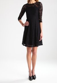 Svart spetsklänning med trekvartsärmar, A-linjeformad siluett, texturerad mönster och knälång fåll. Bärs med svarta stängda skor.