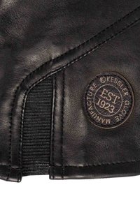 Materiale in pelle nera con superficie texturizzata, caratterizzato da un'etichetta rotonda con "Kessler Glover Manufacture EST 1923". Cuciture visibili e dettaglio elastico.