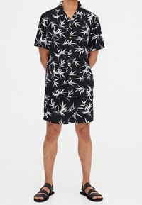 Camisa negra de manga corta y pantalones cortos a juego con un patrón de hojas de bambú blancas, con botones marrones y sandalias negras planas.