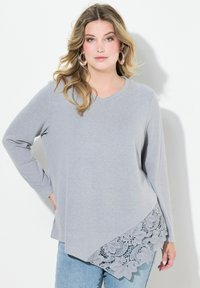 Grå langærmet top med V-udskæring og blonde detaljer ved kanten, der har en asymmetrisk skæring og blødt, tekstureret stof.