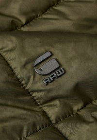 Mörk olivgrön quiltad tyg har en svart, glansig logotyp "RAW" med en distinkt form och en slät textur.