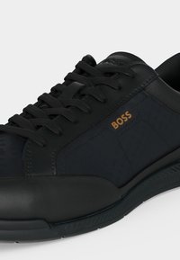 Baskets basses noires avec semelle texturée, lacets noirs, détails en cuir et logo doré "BOSS" sur le panneau latéral.