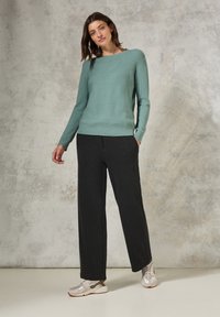 Helles türkisfarbener Strickpulli mit strukturiertem diagonalen Muster, kombiniert mit weiten schwarzen Hosen und grauen Sneakers. Neutraler Hintergrund.
