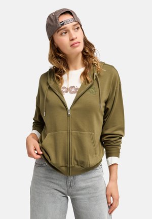 Junge Frau mit einer braunen rückwärts getragenen Kappe trägt einen olivgrünen Zip-Hoodie, ein weißes Blumenhemd und hellgraue Jeans und schaut zur Seite.