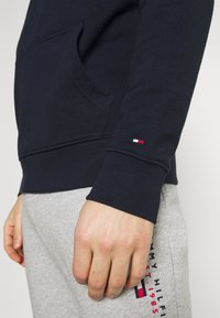 Sudadera de algodón azul marino con un bolsillo frontal, puños acanalados y un pequeño acento de logo rojo, blanco y azul cerca de la muñeca. Combinada con pantalones de chándal grises.