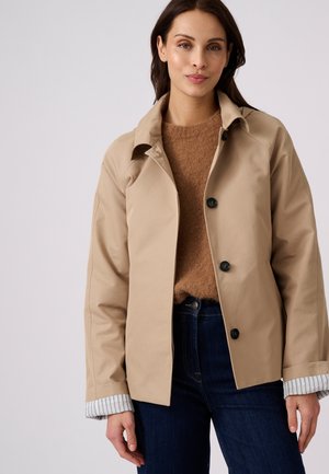 Mujer con chaqueta beige abotonada, jersey marrón y vaqueros azul oscuro, de pie frente a un fondo liso y claro.