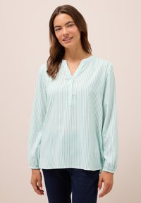 Mujer vistiendo una blusa de manga larga a rayas verticales en azul claro y blanco con botones y jeans azul oscuro, de pie frente a un fondo liso.