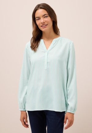 Kvinde iført en lyseblå og hvid stribet langærmet bluse med knapper og mørkeblå jeans, stående foran en ensfarvet baggrund.