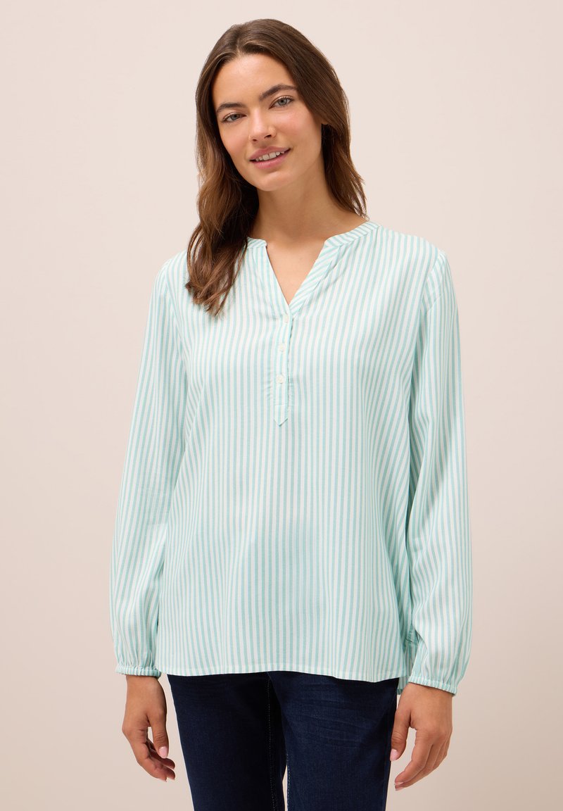 Mujer vistiendo una blusa de manga larga a rayas verticales en azul claro y blanco con botones y jeans azul oscuro, de pie frente a un fondo liso.
