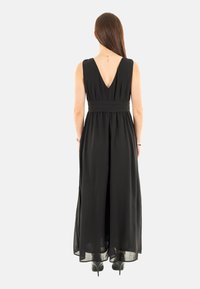 Robe longue noire avec un dos en V profond, taille cintrée, tissu fluide et fentes sur les côtés. Portée avec des chaussures à talons hauts noires.