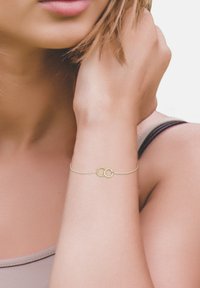 Bracciale in oro con due cerchi intrecciati, uno dei quali è torcigliato per dare texture. Design della catena delicato, indossato su un polso liscio e nudo.