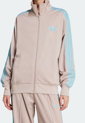 Person iført beige Adidas træningsdragt med lyseblå striber på ærmer og bukser, stående med hænderne afslappet langs siderne.